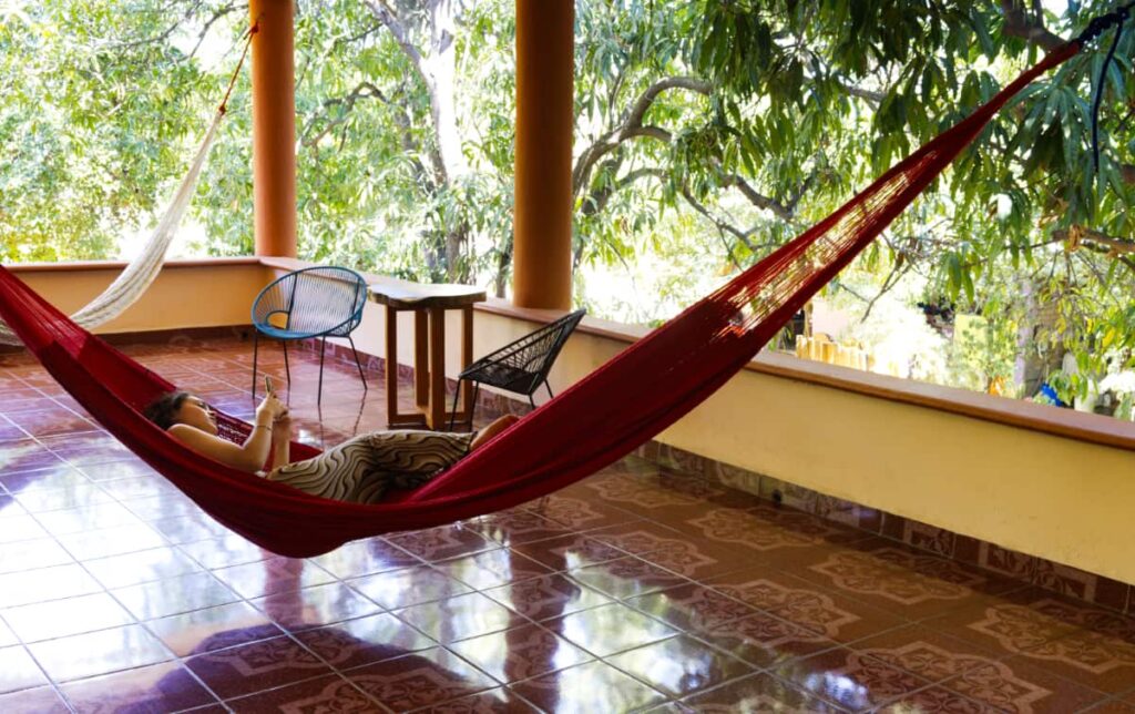 A girl looks at her phone while lounging in a red hammock at Casona Dreams Hostel, Puerto Escondido. Green trees surround the shaded balcony.