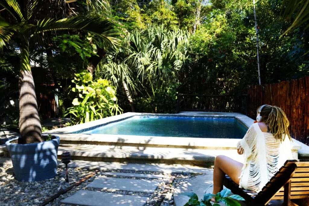 A woman with blonde dreads and headphones lounges by the pool at Selina, Puerto Escondido. The pool is surrounded by green plants and a small potted palm tree.