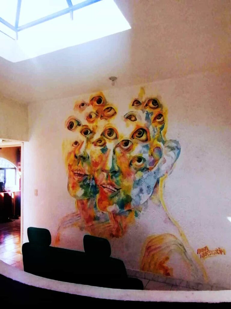 A multi-color wall mural at Vivo Escondido Hostel depicts two male faces attached at the shoulders with many eyeballs looking up towards the skylight.