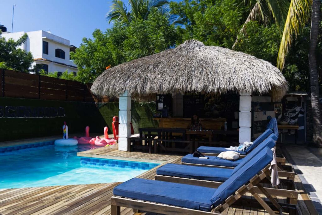 Wooden lounge chairs with blue cushions sit empty in front of the pool and palapa bar area. Bright flamingo and unicorn pool floats sit in the corner of the pool.