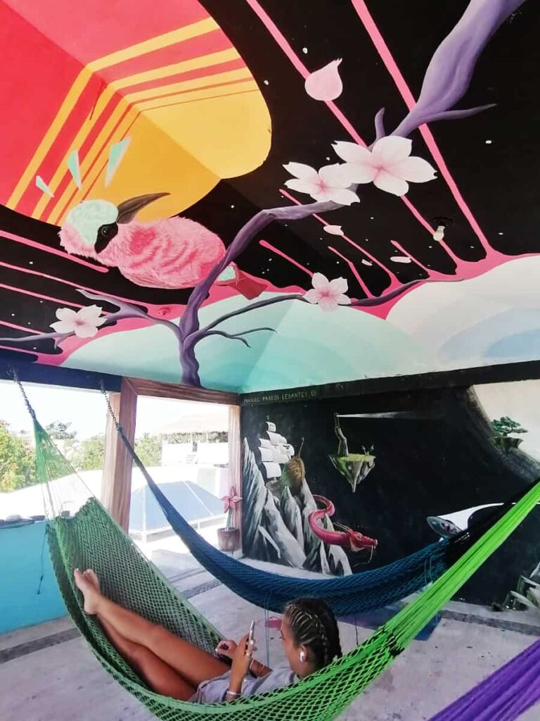 On the rooftop at Vivo Escondido Hostel, a woman lounges in a hammock while looking at her phone. On the ceiling above is a large mural of a bird sitting on a branch with flowers and a large sun behind.
