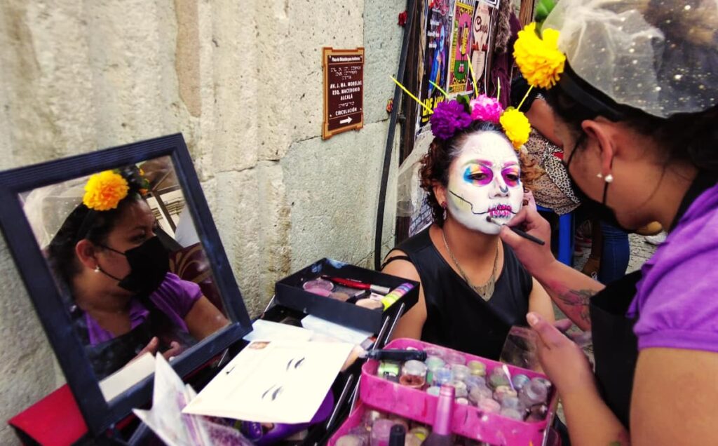 A woman sits to get her Day of the Dead face painted. Her face is all white with a rainbow of blue yellow and pink around her eyes and pink skeleton-like mouth. In her hair is a purple, pink, and yellow flower headband attached to a small white veil.