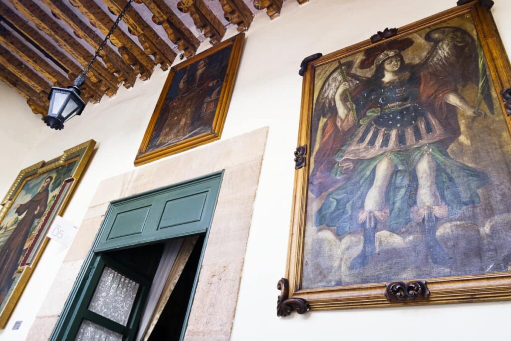 Old pieces of art, mostly religious in nature, hang on the walls of the Queretaro Museo Casa de la Zacatecana. In the center is a open doorway, painted a dark teal with curtains hanging on the other side.