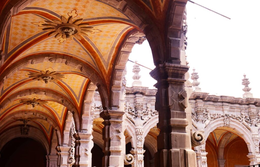 The walkways at the Queretaro Museum of Art features a series of archways with intricately painted details. The patio facade in the background has a series of relief statues of men that served as rain gutters for the building.