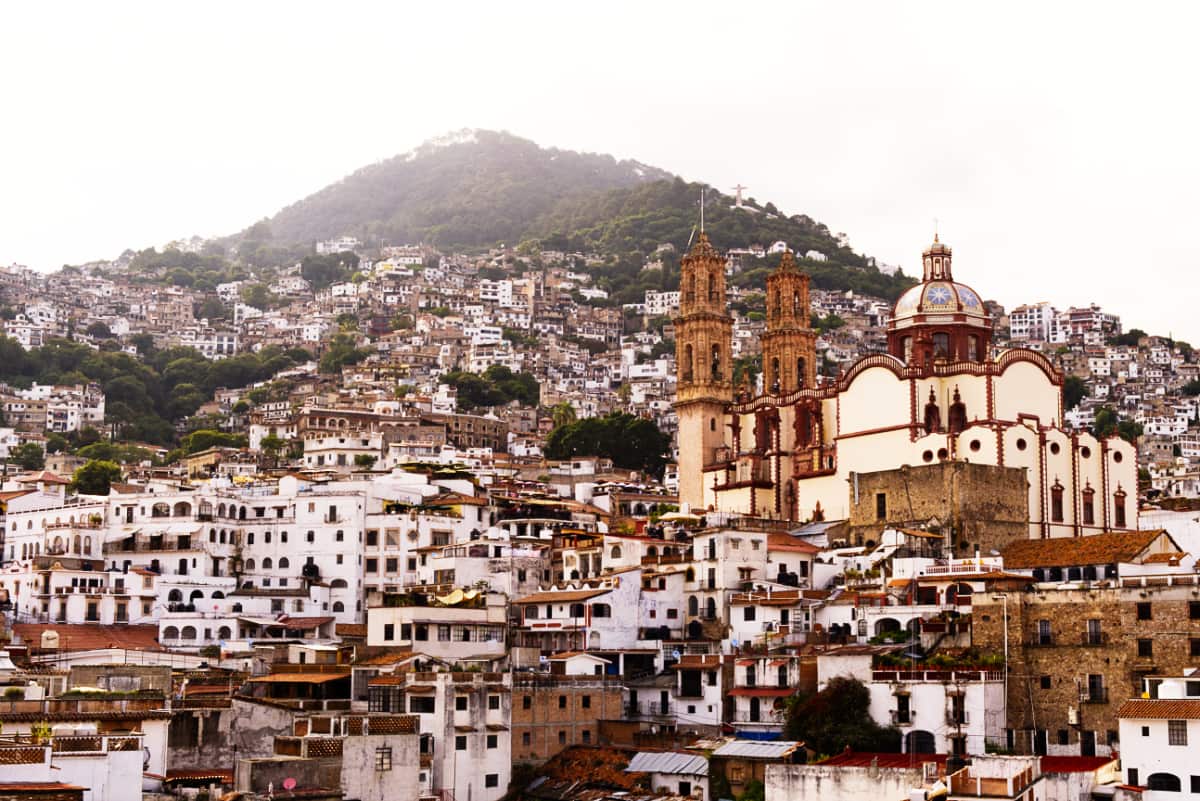 This overall view of Taxco Guerrero Mexico shows the back of the Santa Prisca Church and many houses stacked together into the mountainside. Far in the background is the Monumento Cristo del Rey.