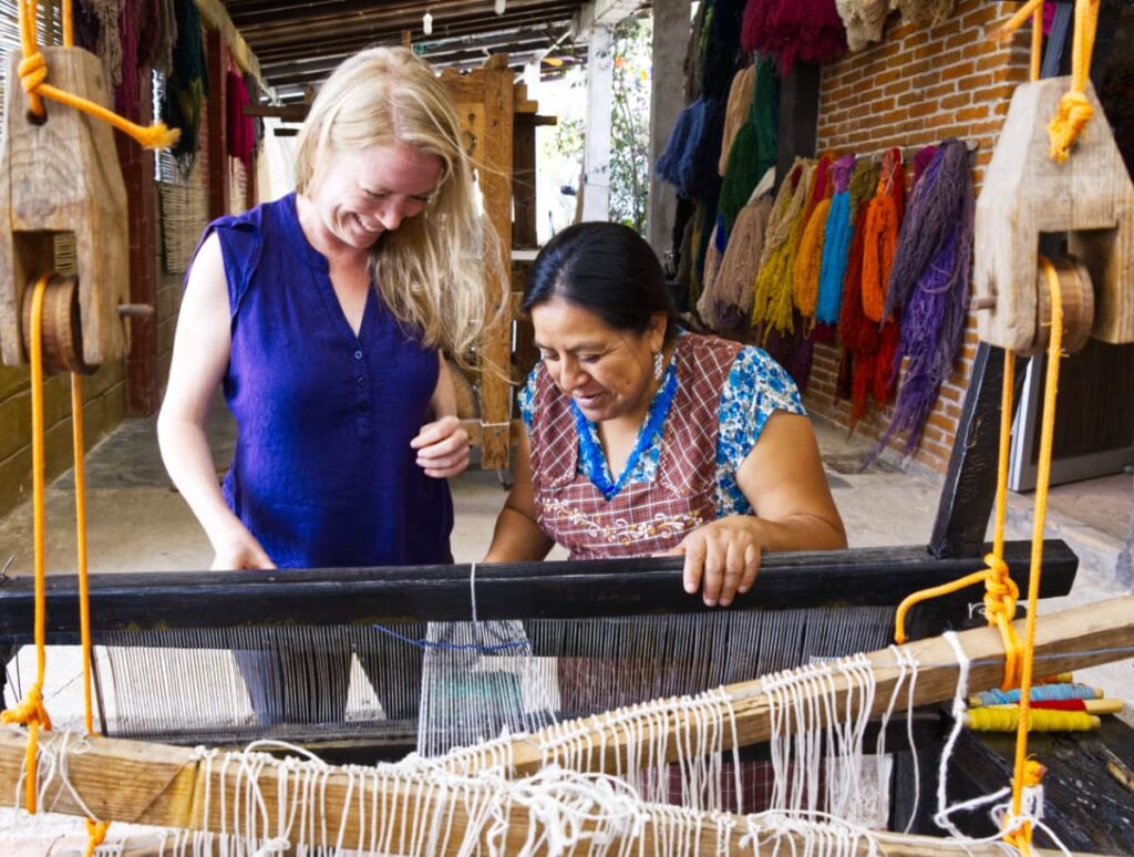 Framed by the 100-year-old loom, we laugh as Josefina helps me with my design during a Teotitlan del Valle weaving tour. Multiple tension threads hang from the wood bars that crisscross in front on the frame.
