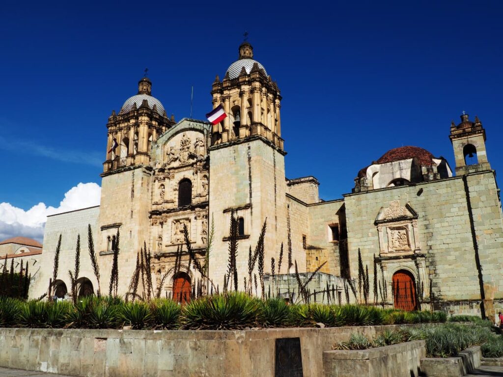 The baroque facade flanked by two towers topped with domes in this outside view of Santo Domingo Church in Oaxaca with succulent plants in the foreground.