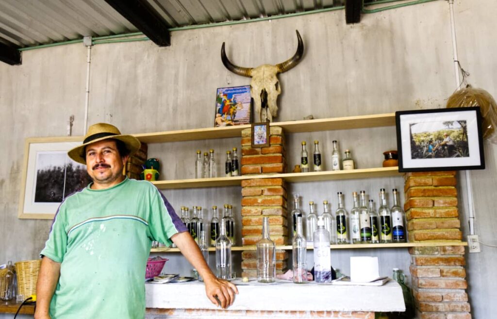 Wearing a hat and bleach stained shirt, Conejo poses in the mezcal tasting room. A skull with horns hangs above the shelves.