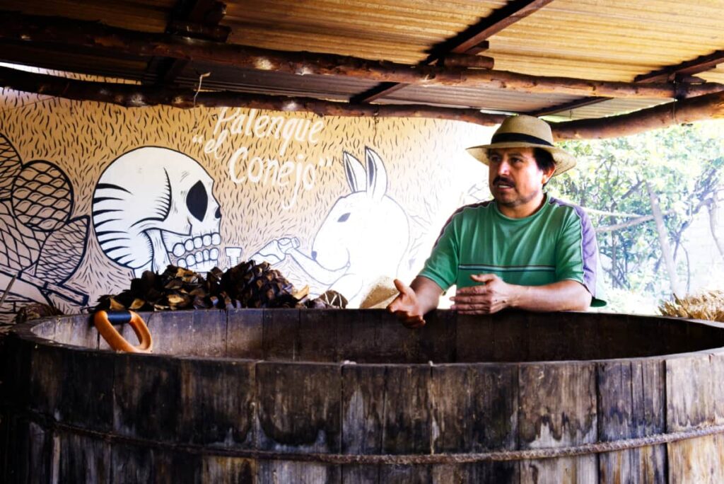 Conejo stands alongside a large, wooden fermentation container with roasted agave hearts piled behind him.