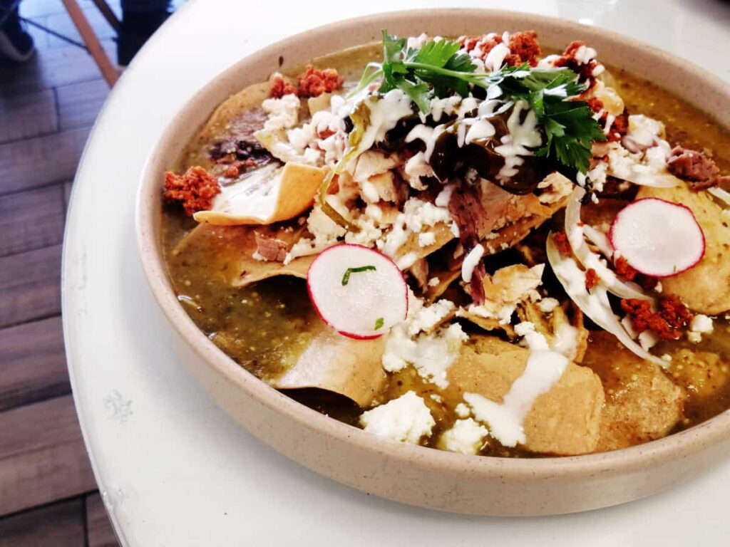 Chilaquiles on a round plate. Tortilla chips are layered with chorizo, cheese, and a green sauce. Topped with sliced radishes and onions.