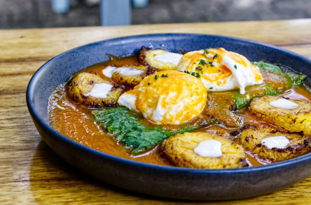 A close up view of one of the best breakfast, Oaxaca. On a round black plate are two poached eggs on top of a stuffed hoja santa leaf which is smothered in a red sauce. Slices of sweet plantain topped with crema are along the side of the plate.