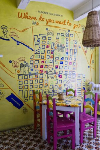 A table and chairs sits in front of a large yellow map of Oaxaca painted on the wall with the words, "Where do you want to go?"