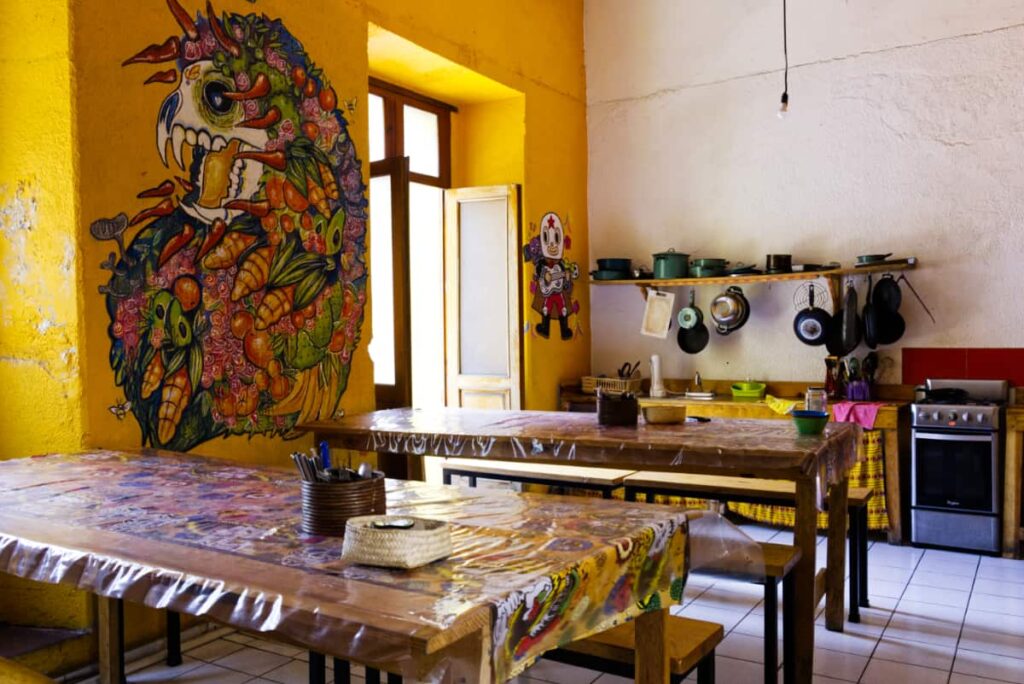 Long wooden tables line the kitchen at Iguana Hostel in Oaxaca. A large mythical animal mural is on the wall and in the back, pots hang above the counter next to the stove.