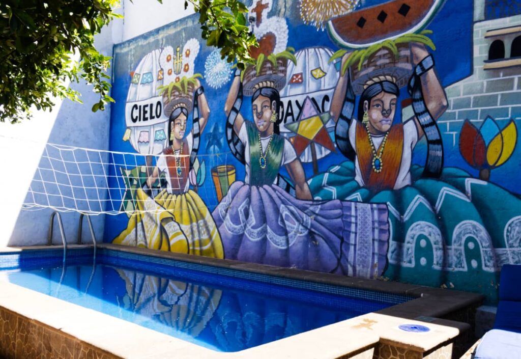A volleyball net is strung across the small rectangular pool at El Cielo Hostel. On the back wall is a mural of three indigenous women carrying baskets on top of their head during a celebratory parade in Oaxaca