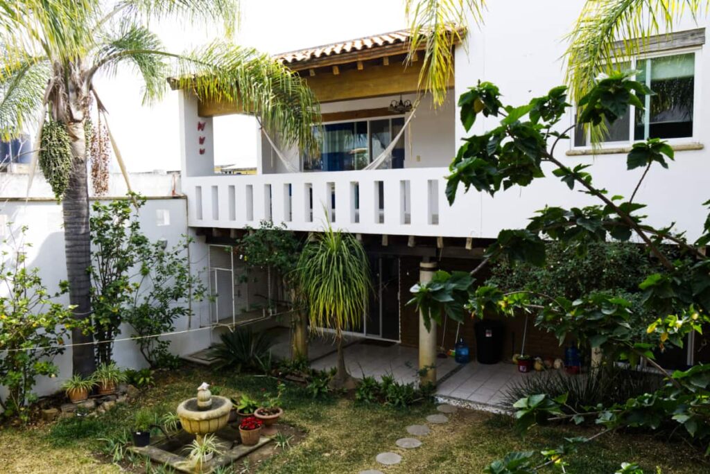 Looking over the back garden at El Jardin Surfista Hostel, plants, a palm tree, and small fountain are in the yard. Above is a white balcony with hammock.