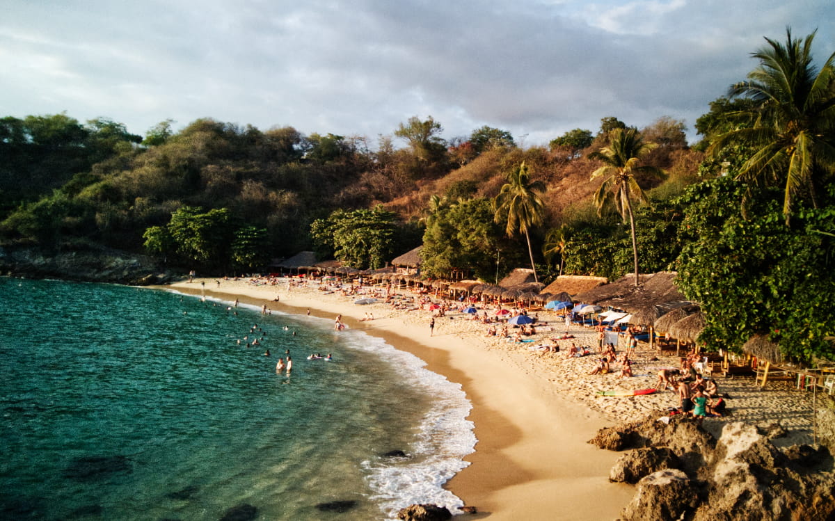 Golden sand and turquoise waters sweep along the bay at Playa Carrizalillo, the most beautiful beach in Puerto Escondido. People lounge in the sand in front of the palapa shaded restaurants while others swim in the ocean.