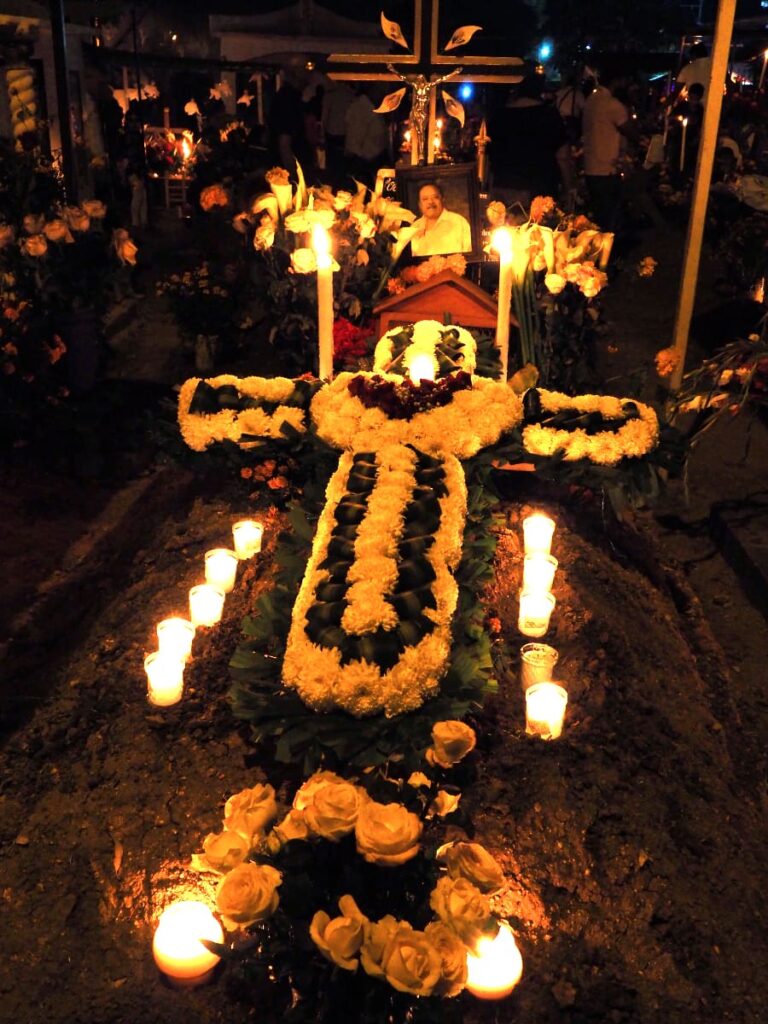 At a cemetery during Day of the Dead in Oaxaca, a gravesite is topped with lit candles, an intricate flower cross, and a picture of the deceased.