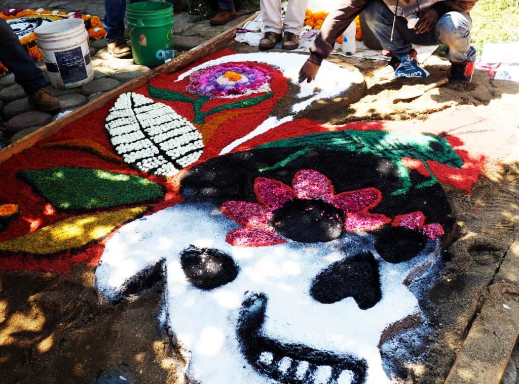 A man squats to work on a sand tapete in Zaachila. In the foreground is a large catrina face with a green grasshopper on top of his head and flowers in the background.