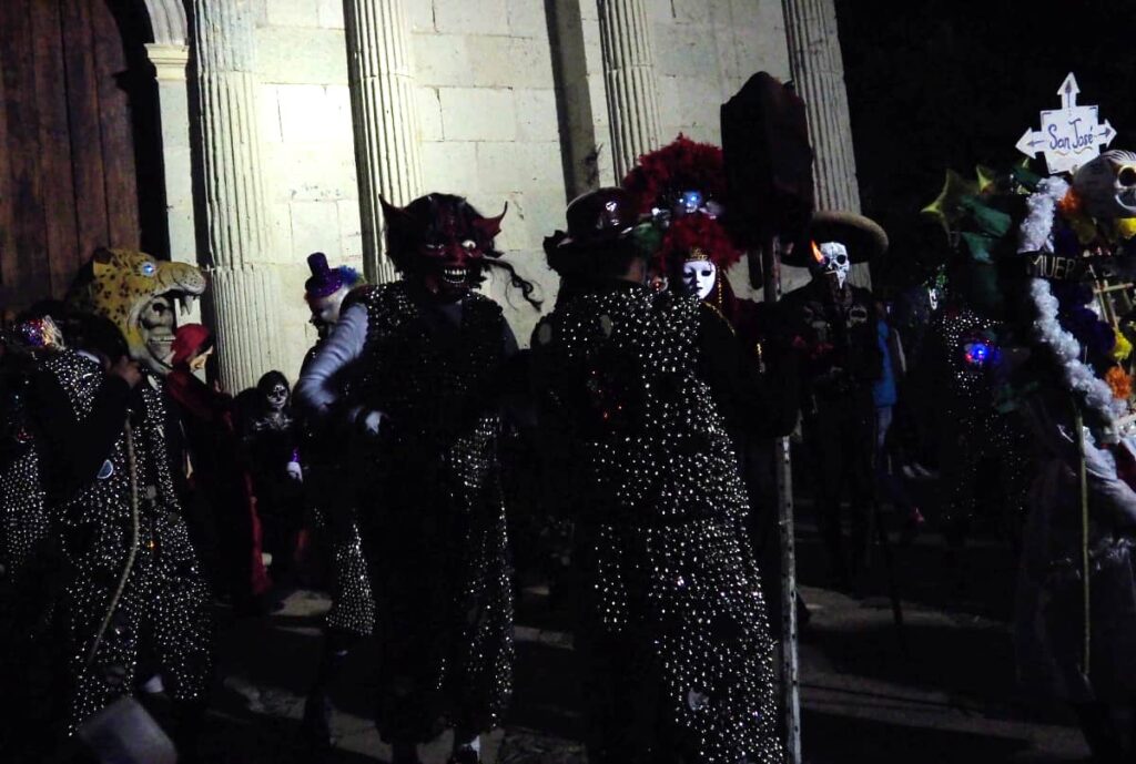 People gather wearing masks and costumes adorned with bells and mirrors in Etla, Oaxaca for Day of the Dead.