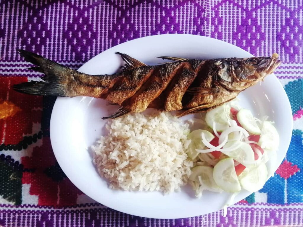 An overview shot of a white oval plate with a whole fried fish, pile of white rice, and a salad of cucumber, tomatoes, and onion. Behind the plate is a woven tablecloth featuring bright patterns in purple, blue, and red.