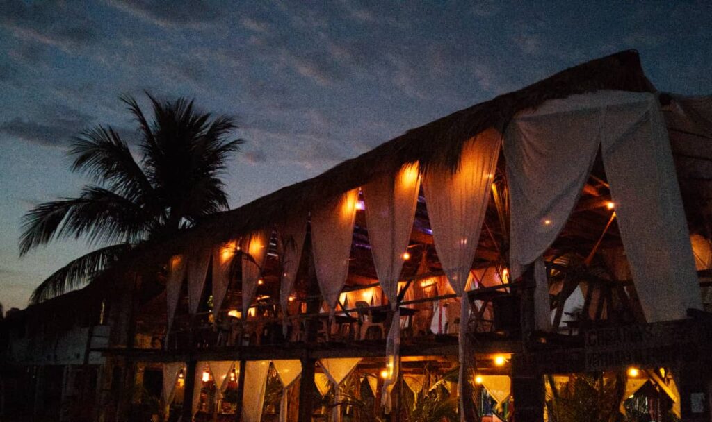 White curtains are draped along the sides of a two story, open air platform on the beach. The artificial lighting creates a club like atmosphere that was not present in Chacahua before. A palm tree is silhouetted against the sky behind the platform.