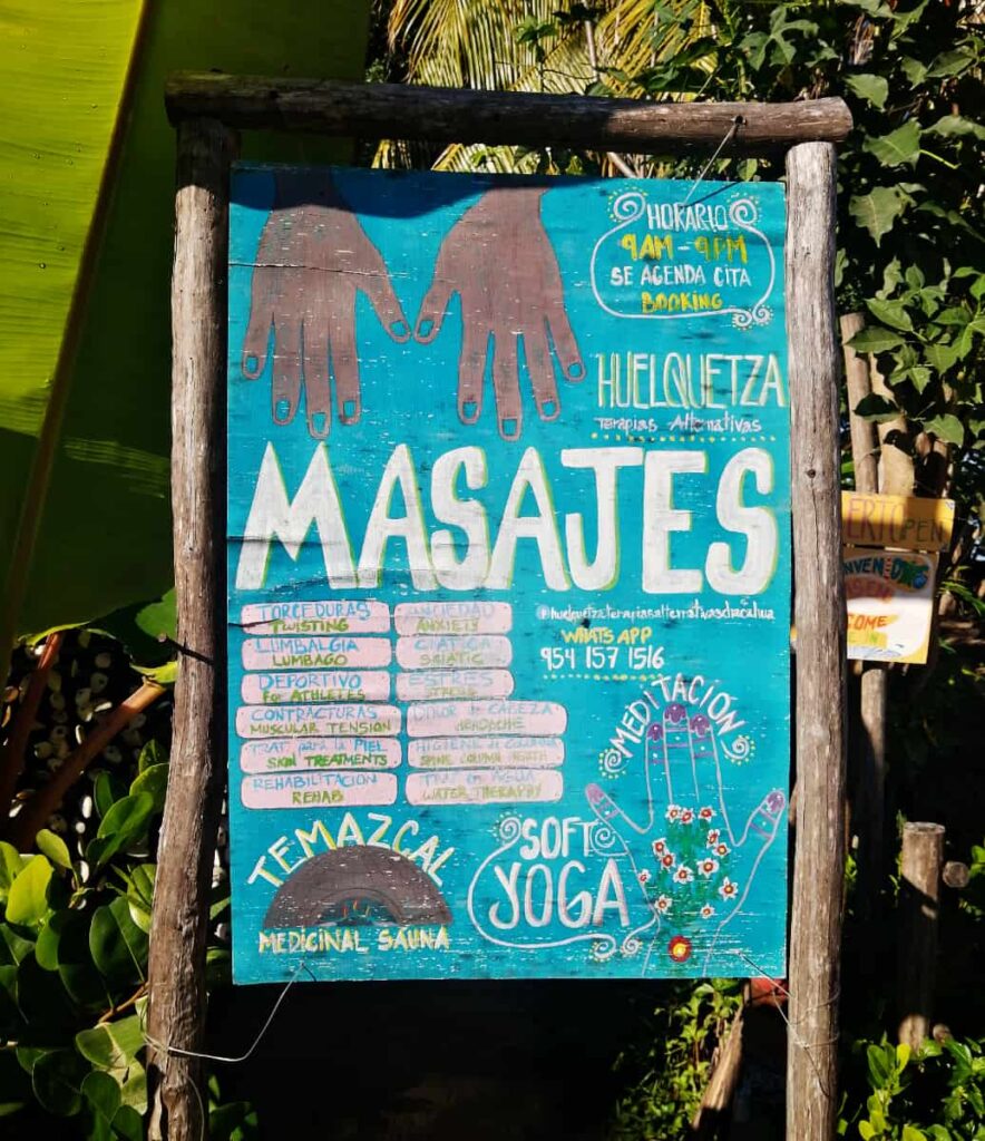 A bright teal, handpainted sign is framed and hung with natural wooden posts. The sign advertises massage, meditation, yoga, temazcal, and other alternative therapies in Chacahua.