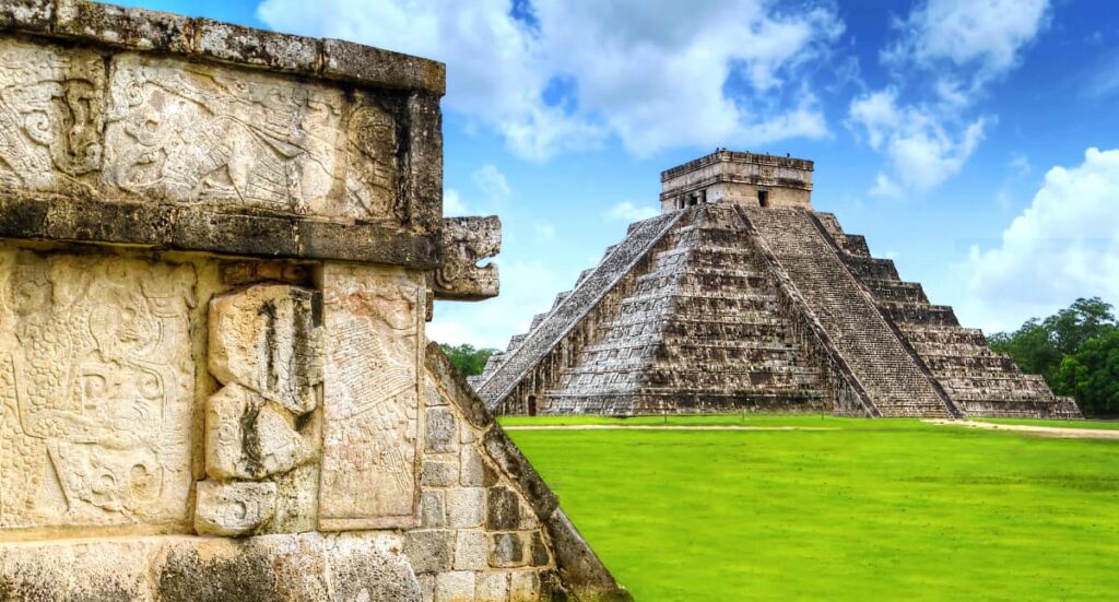 At Chichen Itza Mayan Ruin in the Yucatan, a snake head protrudes from the carvings of one structure. In the background is Kukulkan Mayan pyramid under blue skies with green grass.