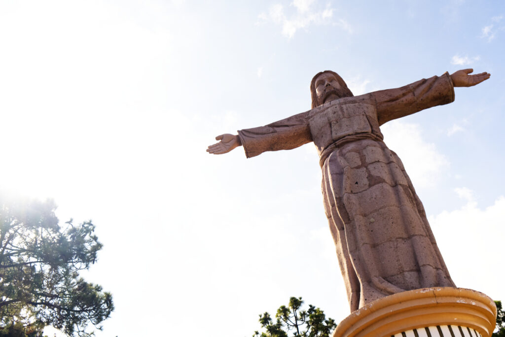 The stone statue of Christ in Taxco, Mexico stands on a small podium and is backlit by the sun.