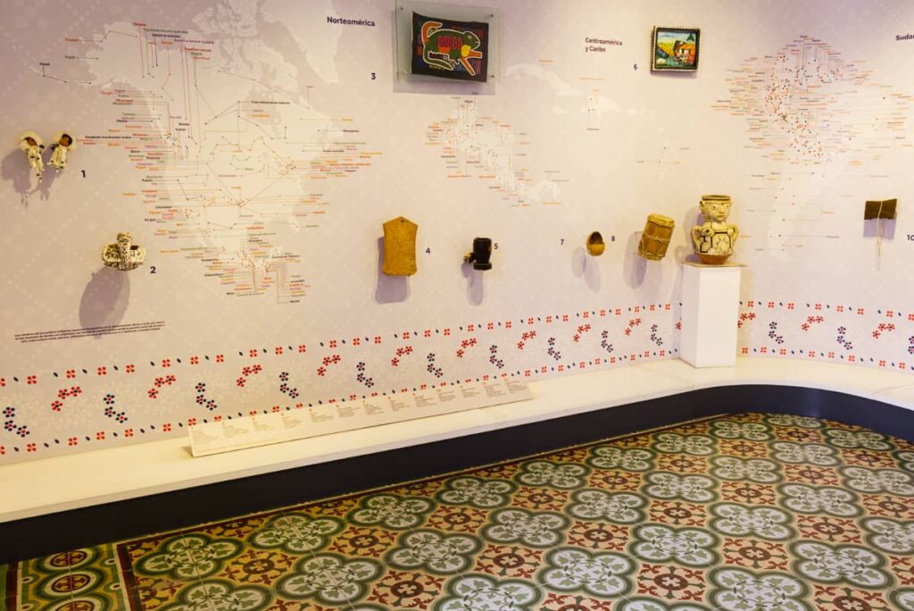 At the Indigenous Museum in Queretaro, a large map of the Americas details various indigenous groups. Clay, stone, and wood artifacts hang on the map wall. On the floor are colorful pasta tiles with a clover pattern.