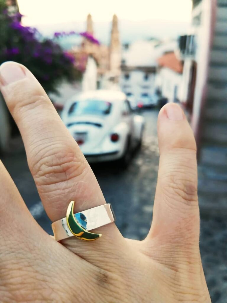 A woman's hand shows off a silver ring with a gold crescent moon. In the blurred background is a classic white taxi of Taxco, Guerrero.
