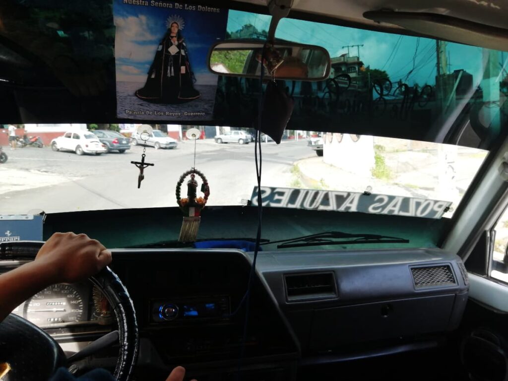 Inside a colectivo van, a man's hand rests on the steering wheel. On the windshield reads Pozas Azules backwards as well as hanging religious art.