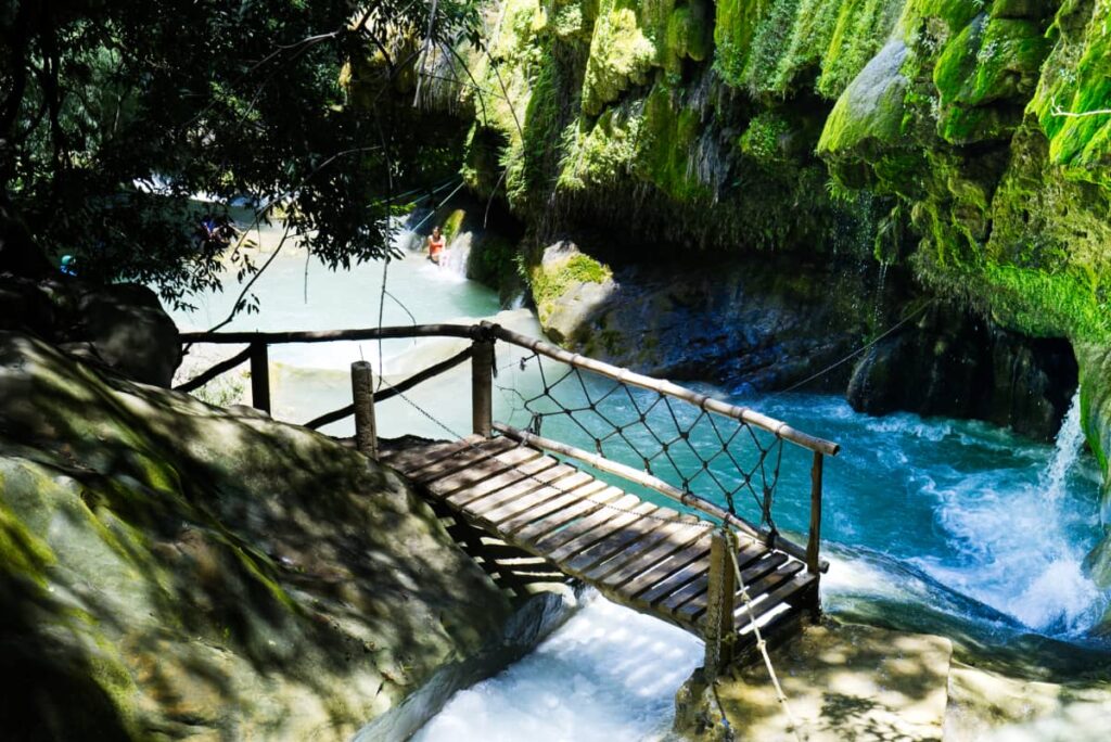 At Pozas Azules, a wooden bridge suspends over a river, between two rocks with a blue pool of water and small waterfall behind. In the background, a woman in a red bikini rests on the side of another blue pool.