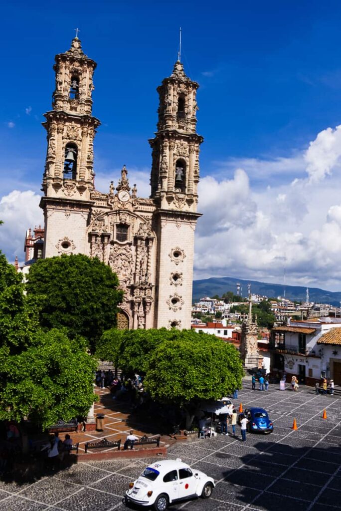 This vertical shot captures the tall columns of the Santa Prisca Church in Taxco, Mexico. In the foreground is Plaza Borda and a white taxi VW bug.