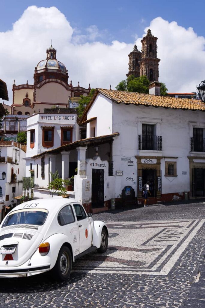 A white VW bug passes in front of the classic Taxco Hotel, Los Arcos. Most of the buildings are white and the street is paved in cobblestones with the year 1967.