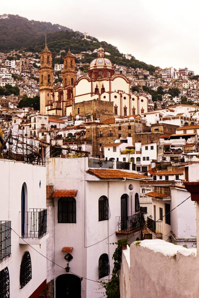 In this vertical shot, white houses are stacked together with the Santa Prisca Church and mountains in the background.