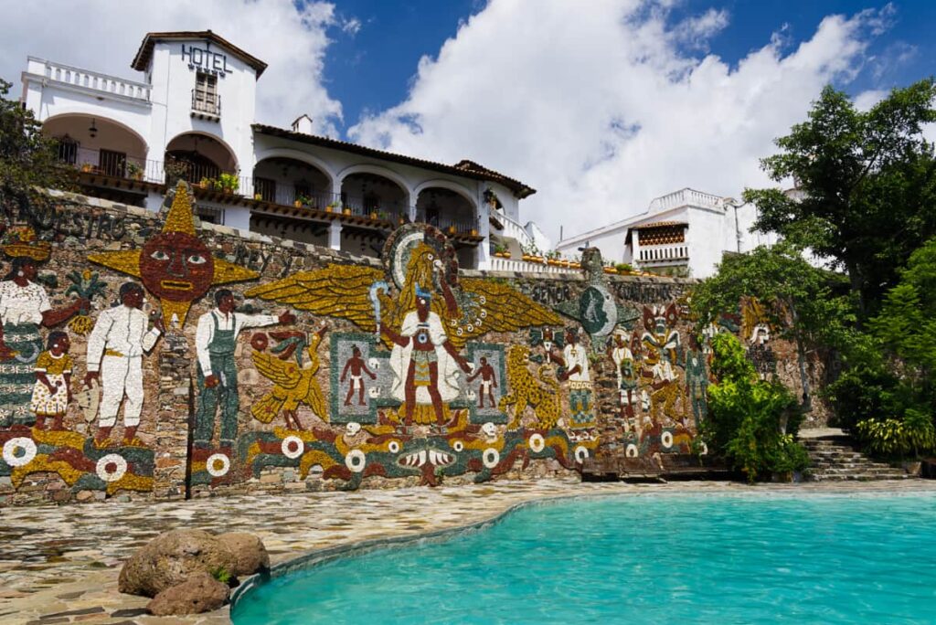 At the preHispanic Silver Mine in Taxco, the bright turquoise pool glistens in the sun with a large indigenous mosaic adorning the back wall. Behind the pool is another white building that says Hotel.