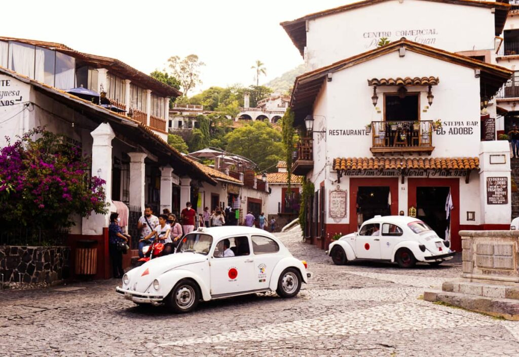 Two white VW bugs, the taxis of Taxco, Guerrero traverse through the cobblestone streets. In the background are white buildings with terracotta roofs.