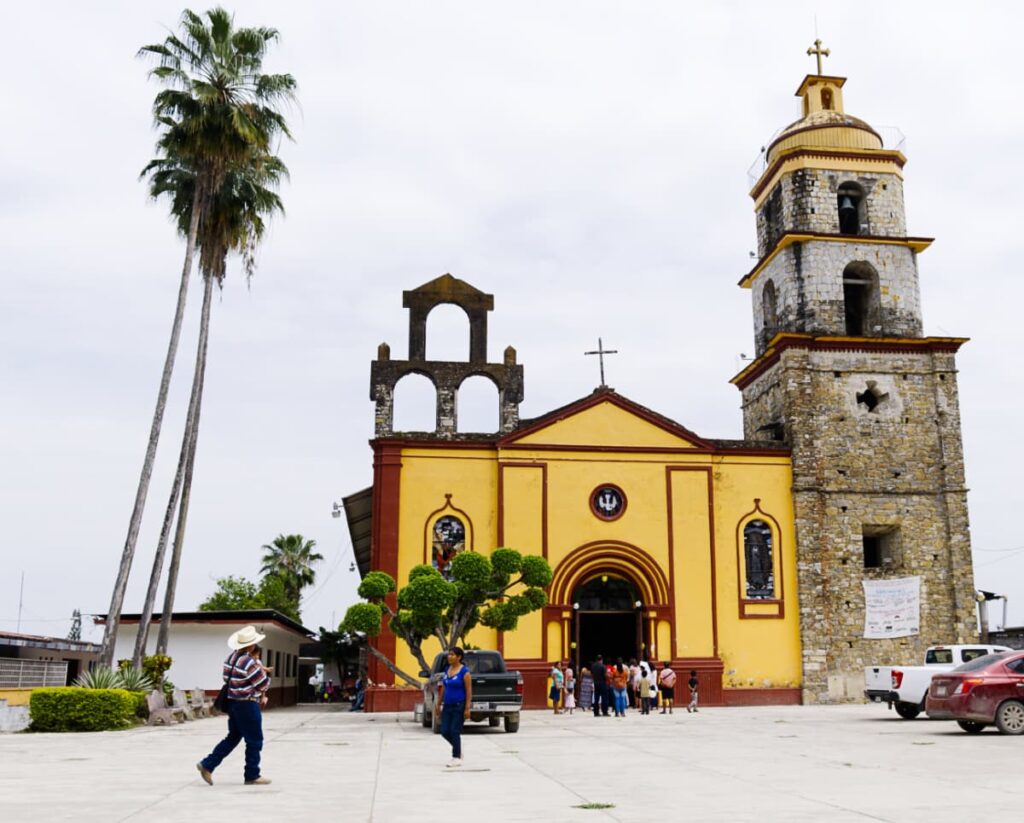 In front of the yellow church in Aquismon, San Luis Potosi a man in a white cowboy hat walks towards a woman. On the left is a tall palm tree. The yellow church has red trim and a bell tower on the right with a cross.