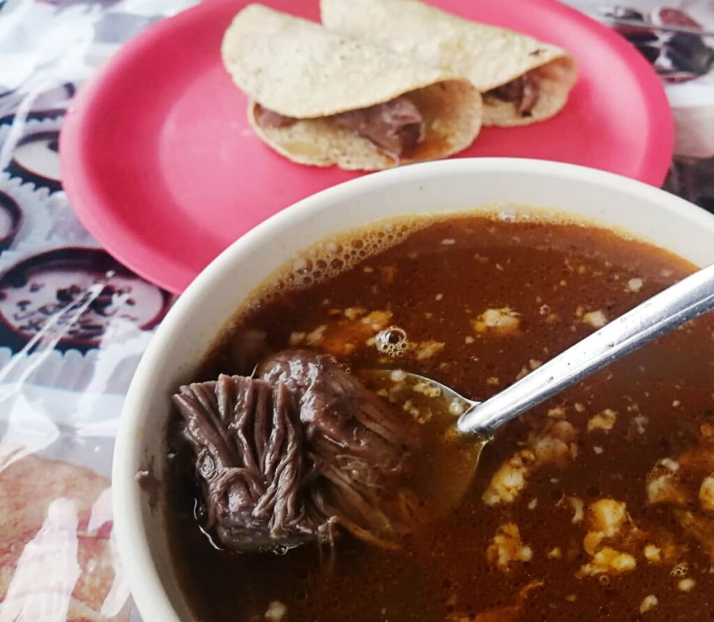 A spoon lifts lamb meat to the surface of a bowl of consume in Ciudad Valles, Mexico. In the background is a red plate with two barbacoa de borrego tacos.