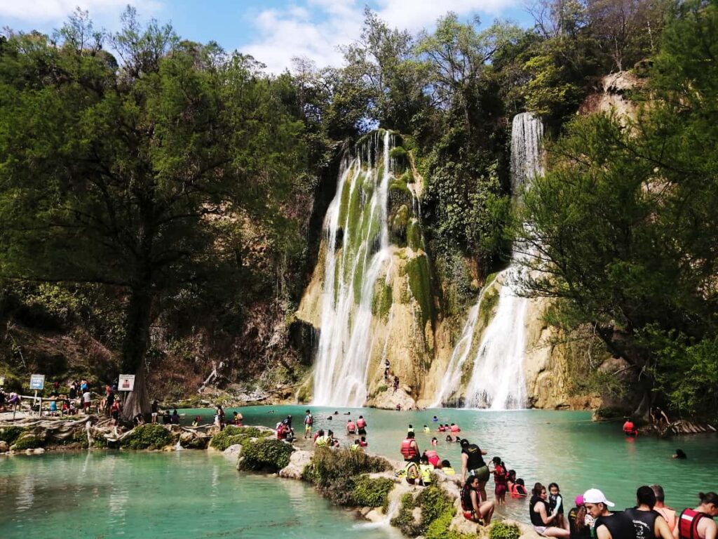 In front of the Minas Viejas Waterfalls in San Luis Potosi, men, women, and children wearing life jackets sit on the limestone bank surrounded by turquoise water. One of the best Huasteca Potosina tours involves rappeling down the rock next to these waterfalls.