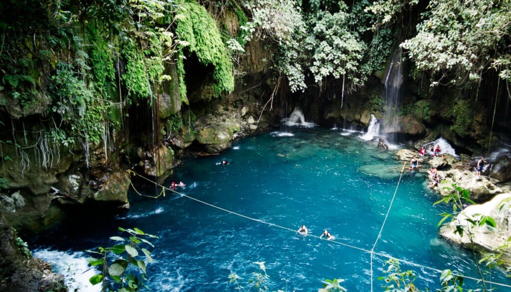 At Puente de Dios in Huasteca Potosina, Mexico, two girls swim in a blue swimming hole while holding onto a rope. Surrounding the swimming hole are rocks with lush vegetation, small waterfalls, and other people swimming.