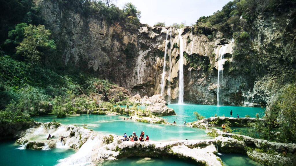 Water cascades from Salto del Agua, Huasteca Potosina into the turquoise pools below. People stand on the banks of the pools while others swim.