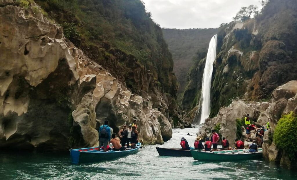 In front of the Tamul Waterfall, San Luis Potosina, people wearing life jackets stand and sit in wooden boats. The try to get a glimpse of Cascada Tamul while protected behind large rock boulders in the water.