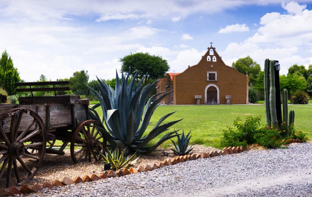 An old hacienda estate is one of the many places you can explore on a day trip from San Miguel de Allende. In this image, a cactus and large maguey plant frame a small church. On the left is an old wooden wagon.