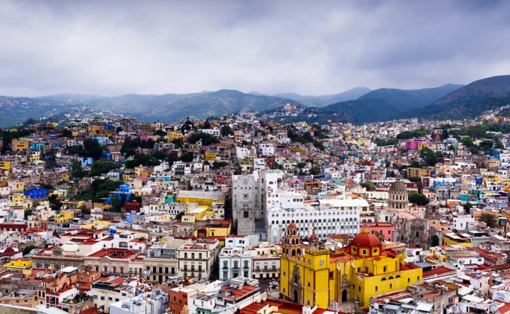A view of Guanajuato, one of the most beautiful cities in Mexico, from the Pipila viewpoint shows many colorful buildings stacked into the surrounding mountains as well as the main church in a bright yellow with red trip.