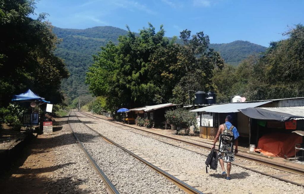 A man wearing a backpack and holding a life vest in his hand walks along the railroad tracks on the way to Puente de Dios, San Luis Potosi. In the background are green mountains. And on the sides are various tents that sell food and drinks.