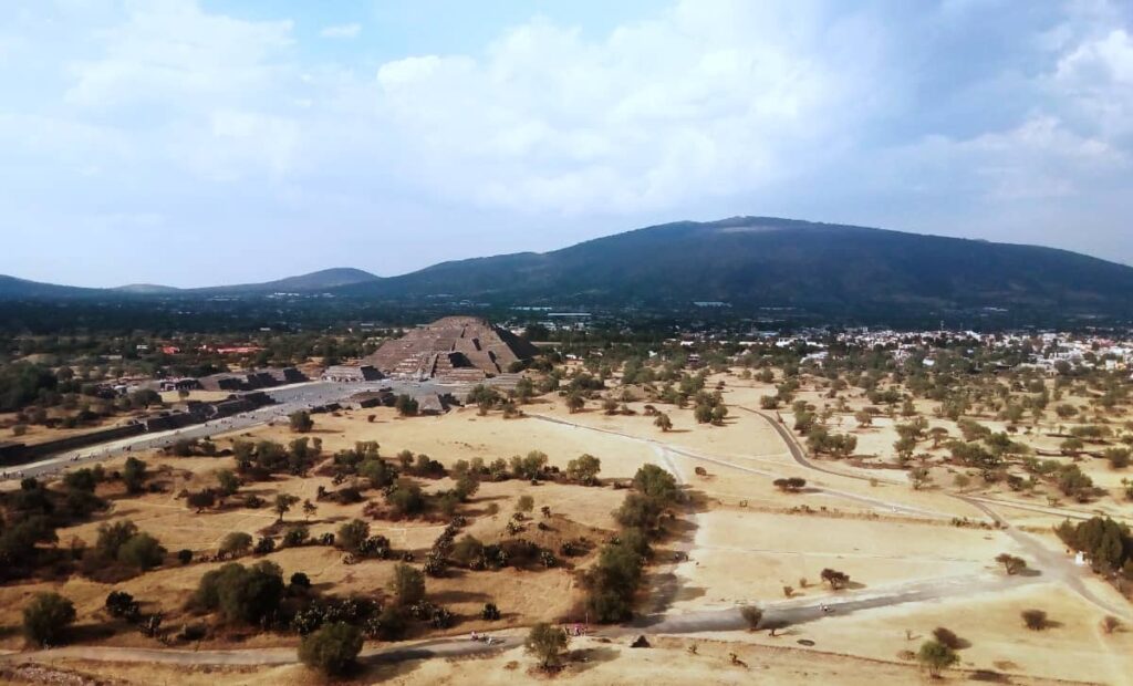 An overview of Teotihuacan, Pyramid of the Moon, and the surrounding mountains. In the foreground is a large field with roads cut in and some small trees.