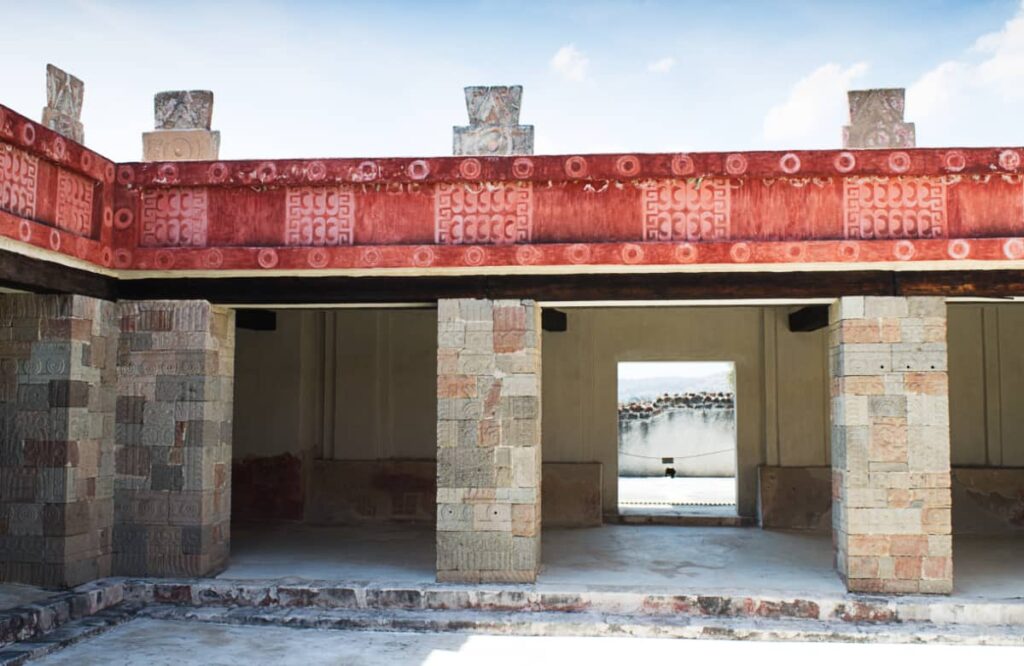 At the Palace of Quetzalpapalotl, Teotihuacan are carved stone columns that lead to a horizontal border with abstract decoration primarily in a red color.