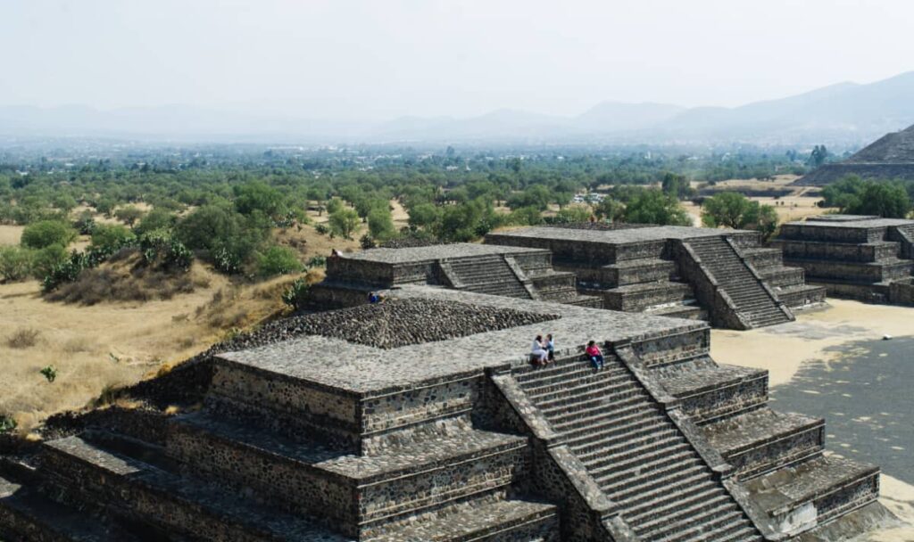 Three people that are visiting Teotihuacan sit at the top of the steps of a small pyramid. In the background are other Teotihuacan pyramids, small trees, and mountains.