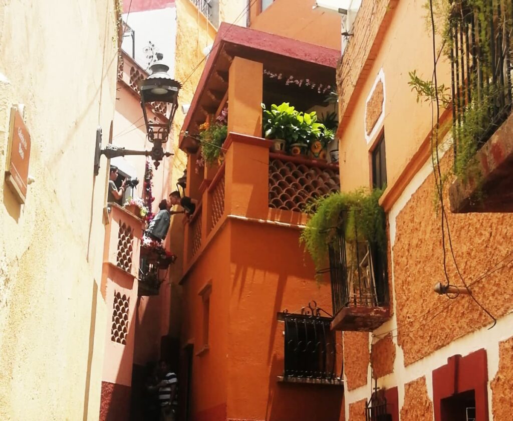 Along the alley of the kiss in Guanajuato, Mexico, a young man and woman lean over the balconies to kiss while a photographer takes their picture. The narrow alleyway is lined with orange colored buildings close together.